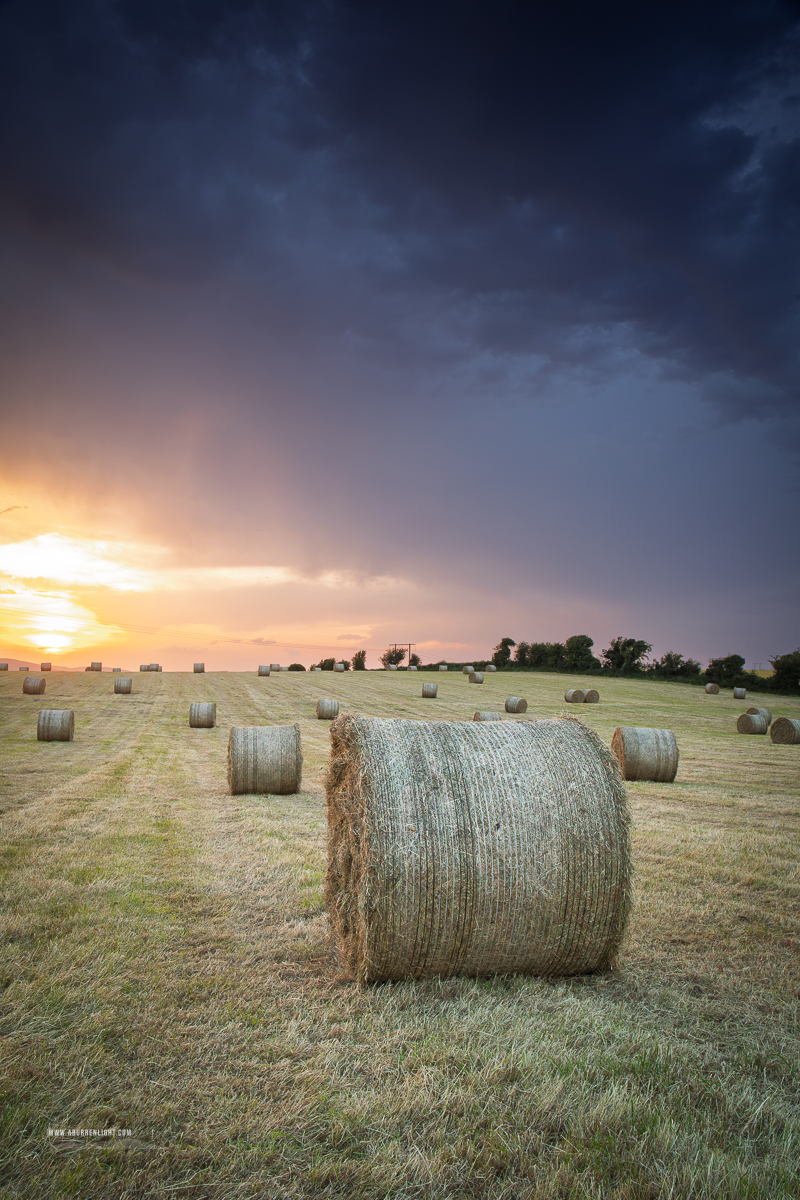 Hay bale beneath dark storm clouds glowing with warm sunset light near Gort, County Galway.