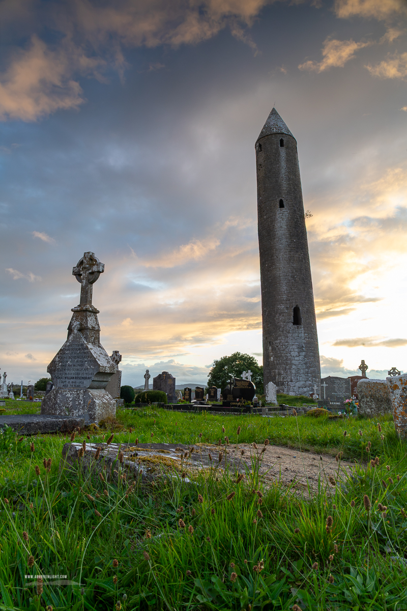 The tall round tower and a Celtic cross at Kilmacduagh Monastery beneath broken clouds and warm evening light.