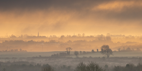 Golden sunrise light filtering through mist across layered fields and trees near Gort, County Galway.