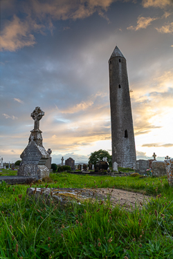 The tall round tower and a Celtic cross at Kilmacduagh Monastery beneath broken clouds and warm evening light.